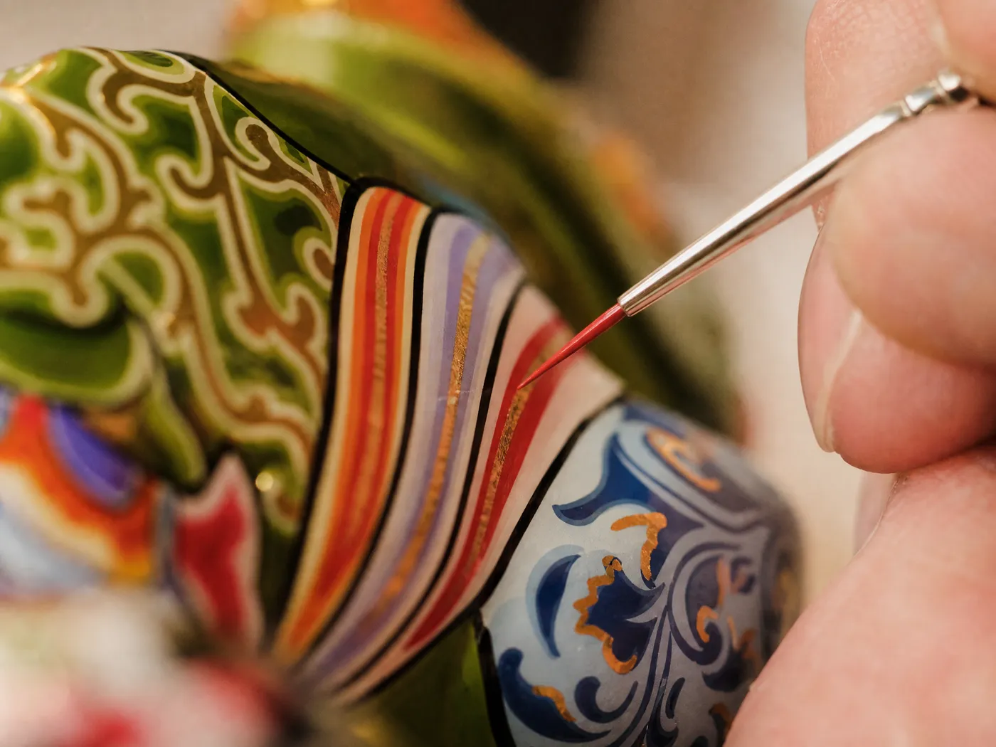 An artisan's fingers painting a fine red line onto a hand-painted chameleon